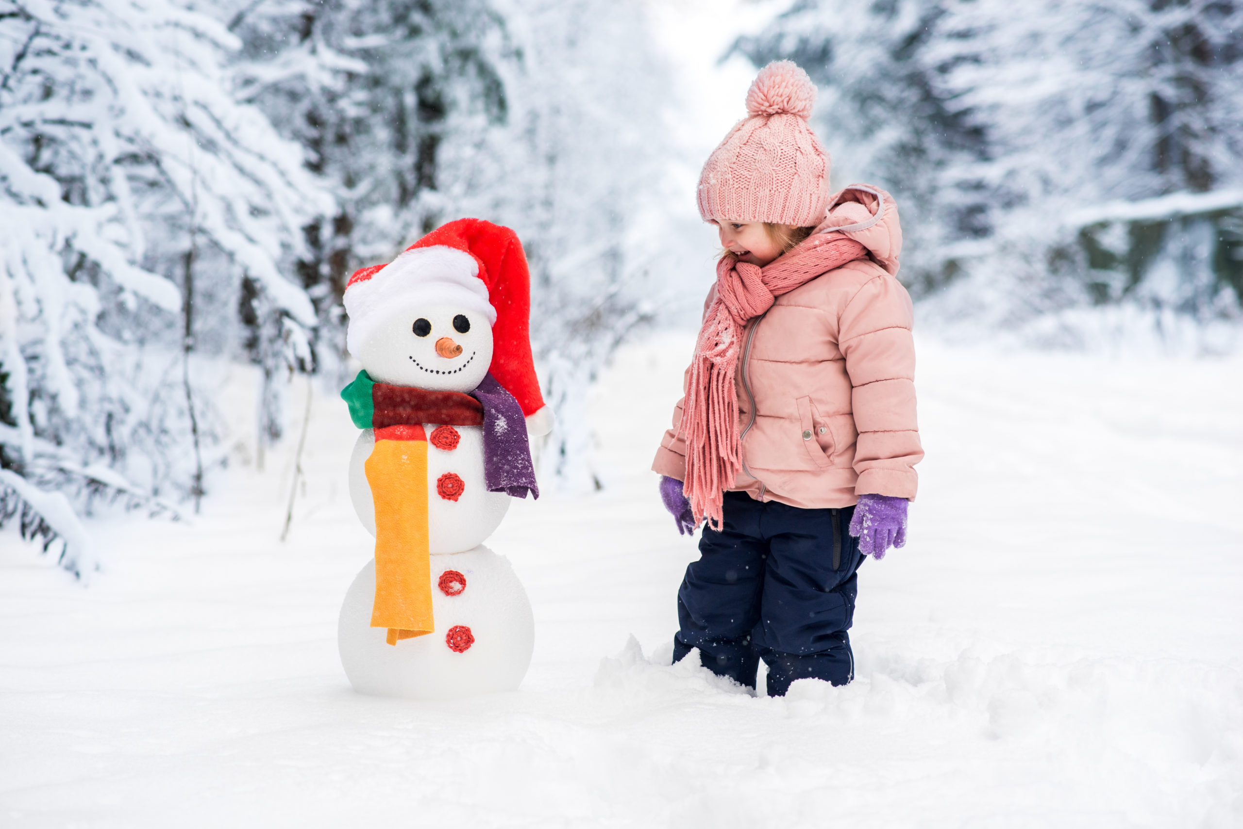 little girl in pink coat and hat smiling at a cute snowman in colourful scarf